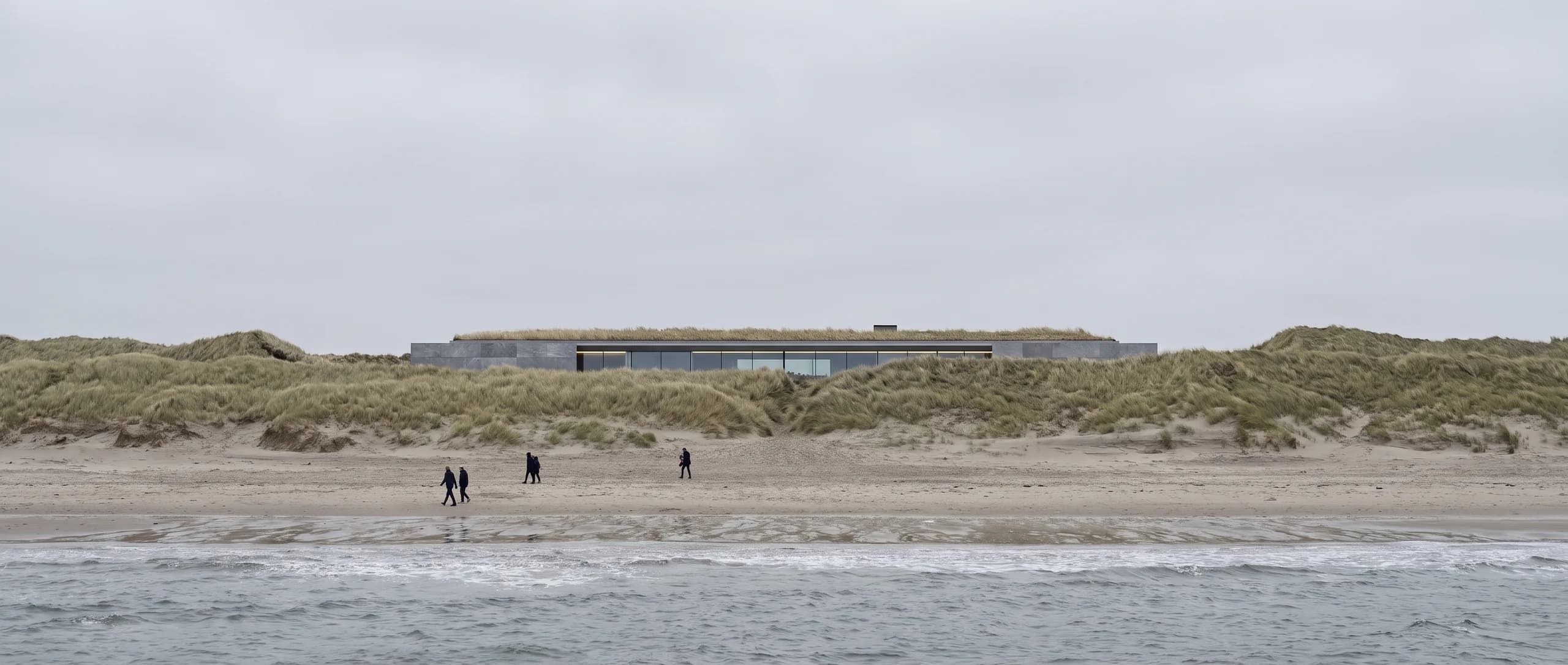 Neutral daylight concept view from the public beach in flat overcast light, showing the low grass-roofed building sitting behind and below the natural dune ridge.